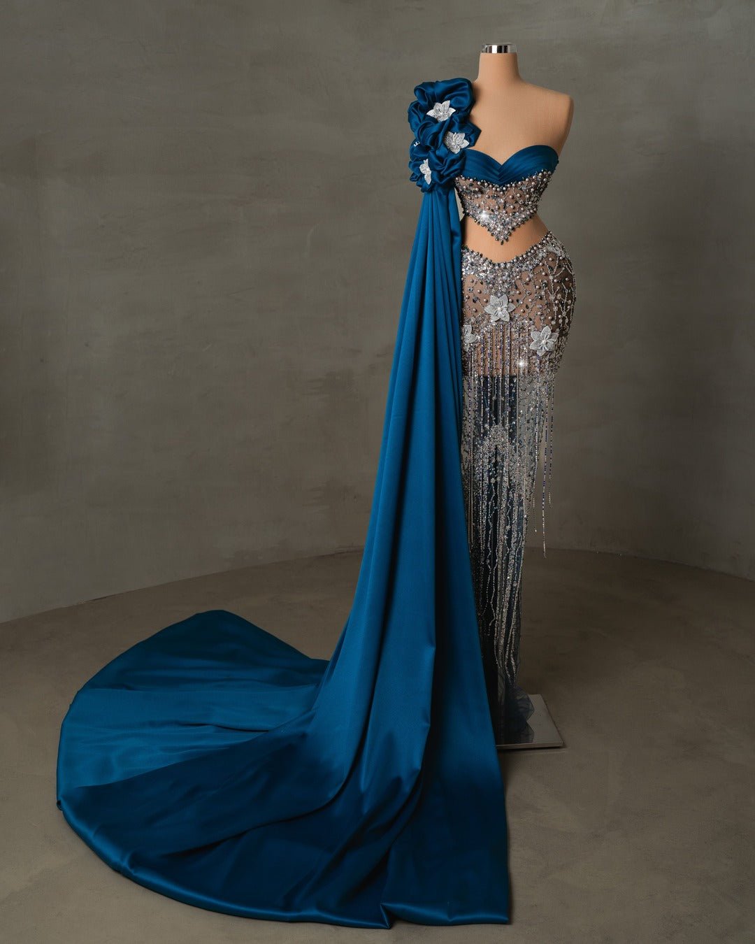 Elegant blue gown with crystal and floral accents, one-shoulder drape, and sheer details along the bodice.